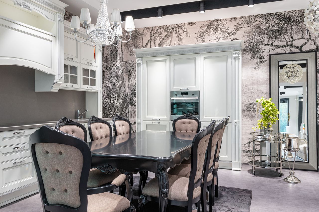 Chairs at table placed near white cupboards and modern oven in spacious stylish kitchen with green plant on side table near mirror
